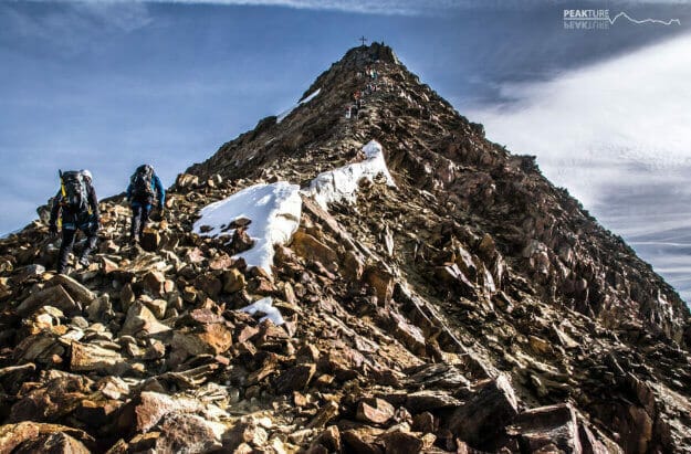 zwei Bergsteiger im schroffen Gelände. Im Hintergrund ist das Gipfelkreuz am Ende des leicht verschneiten Bergrückens zu sehen. Der Himmel ist leicht bewölkt.