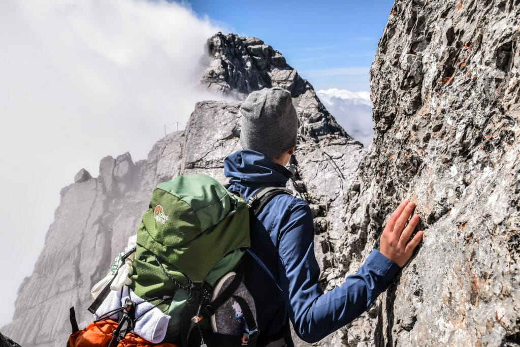 Frau beim Wandern vor einem Zustieg zu einem Alpinen Steig. Im Hintergrund ist ein sehr steiler und ausgesetzter Grad zu sehen