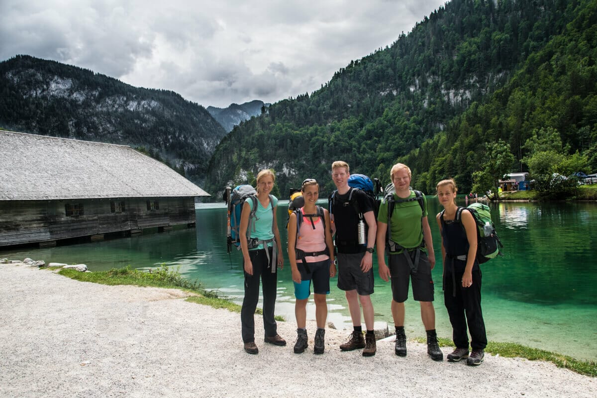 Gruppenbild mit 5 Personen am Königssee im Sommer. Links befindet sich ein Bootshaus und im Hintergrund sind Berge zu sehen