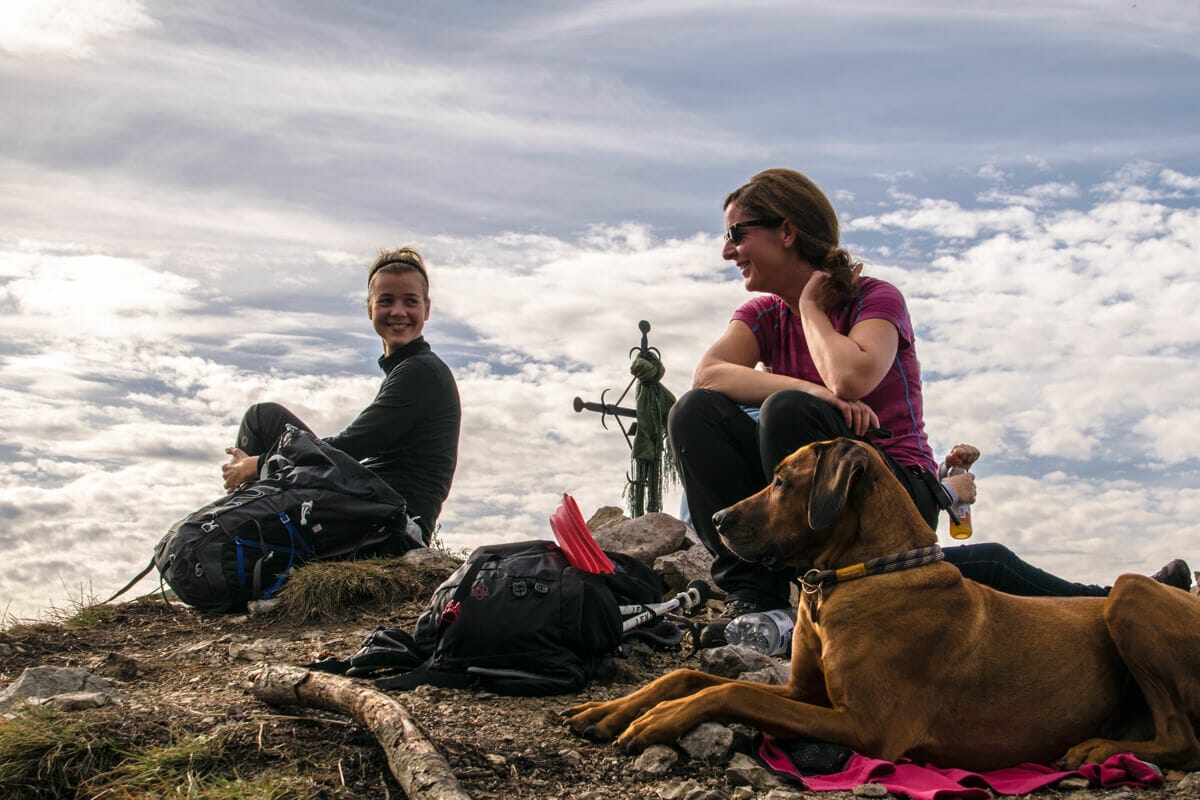 zwei Frauen mit Hund am Gipfel der Sonnenspitze, im Hintergrund Wolken.
