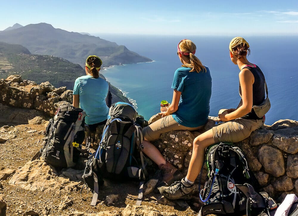 drei hübsche Frauen bei einer kurzen Rast auf dem Trockenmauerweg von Mallorca, im Hintergrund das Meer