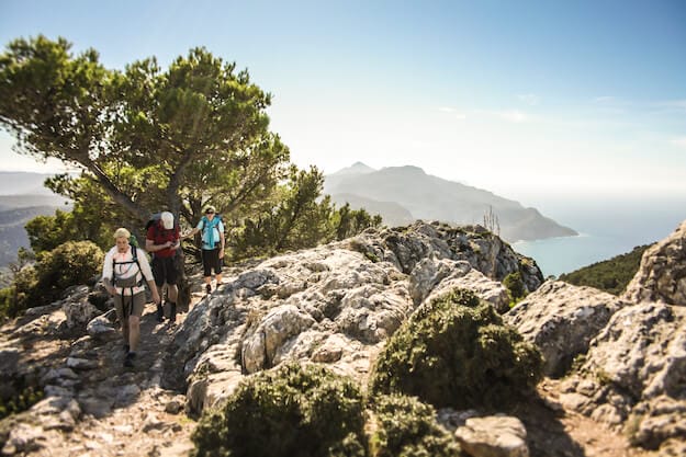 drei Bergsteiger auf einer Wanderung an der Westküste auf Mallorca. Bäume, Felsen und das Meer schmücken den Hintergrund